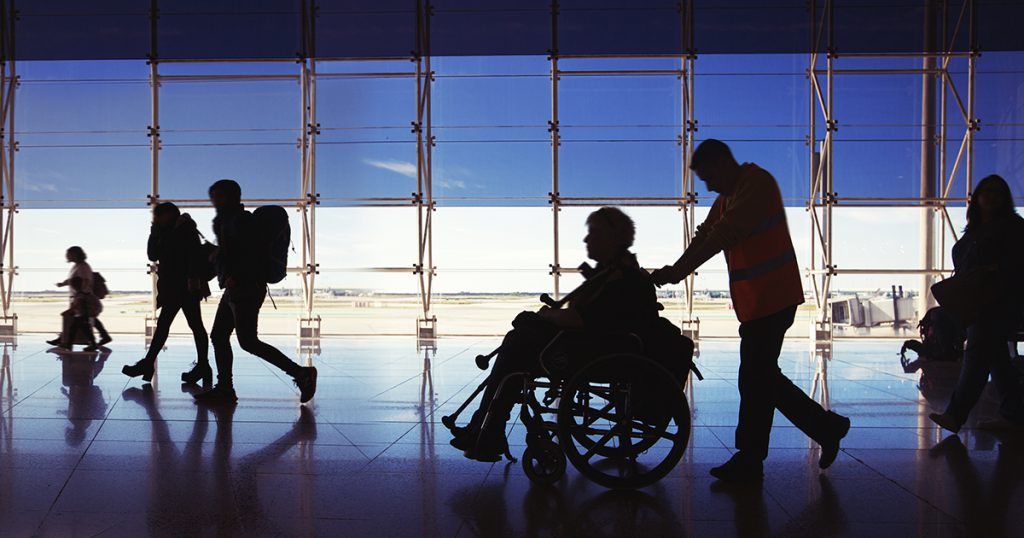 Silhouette of travelers going through the airport, specifically a younger man pushing an older woman in a wheelchair.