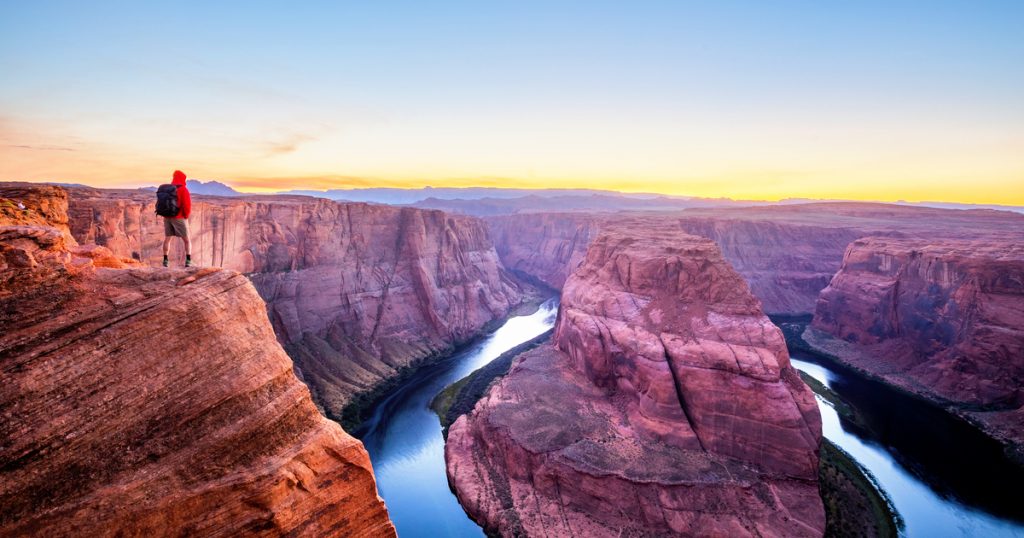A male hiker is standing on top of steep cliffs enjoying the beautiful view of Colorado river flowing at famous Horseshoe Bend in beautiful post sunset twilight on a summer evening, Arizona, USA