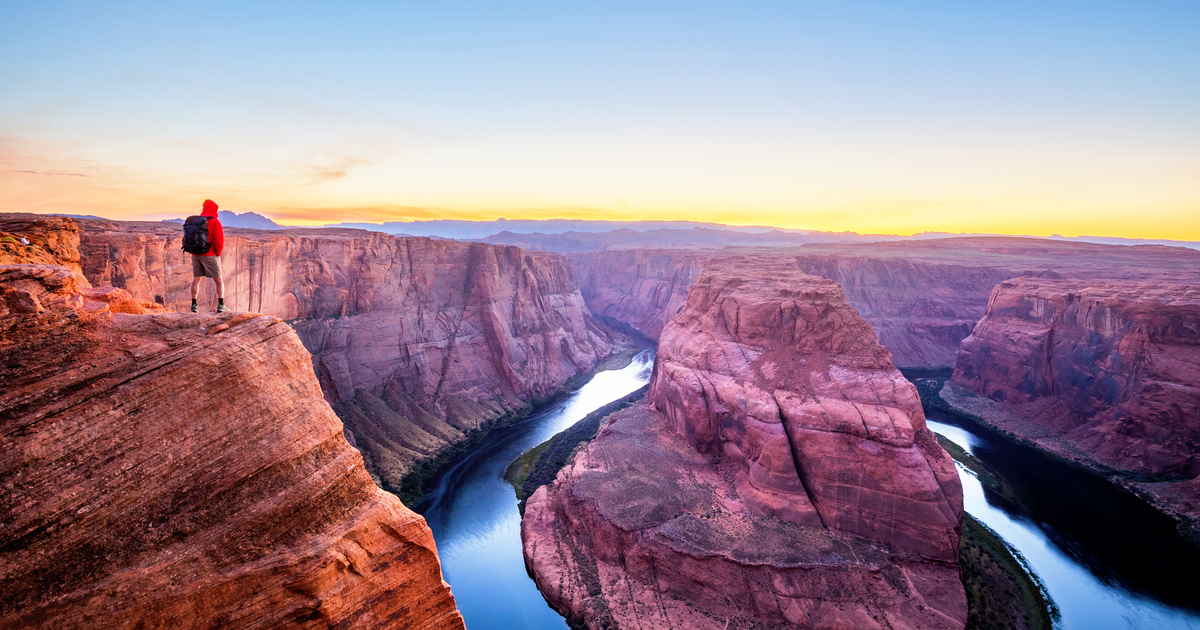 A male hiker is standing on top of steep cliffs enjoying the beautiful view of Colorado river flowing at famous Horseshoe Bend in beautiful post sunset twilight on a summer evening, Arizona, USA